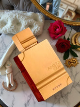A gold and red Al Haramain Amber Oud Rouge Eau de Parfum bottle from Showlu Well Nation Store sits on a marble surface, surrounded by two red roses, a pearl hair clip, a jeweled pin, and a gold mirror.