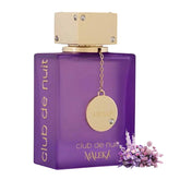 A purple bottle of Showlu Well Nation Store's Armaf Club De Nuit Maleka Pheromone Fragrance for women, with a gold cap and "club de nuit" on the side, is displayed alongside purple flowers—designed to attract the opposite sex.