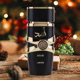A black and gold Lattafa Asad Eau De Parfum bottle (3.4oz) from Showlu Well Nation Store rests on wood with festive lights, pine, and berries—a long-lasting unisex Arabian fragrance, perfect as a gift for any season.