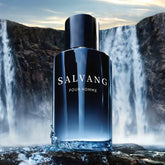 A 3.4 Oz men’s Eau De Toilette spray by Showlu Well Nation Store, with notes of Calabria lemon, pepper, and vetiver, stands in water near a dramatic waterfall under a cloudy sky—perfect for daily use or dating.