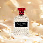 A clear, textured glass perfume bottle with a red cap and black label reading VALENTINE ROUGE sits on golden fabric with gold flakes—this 3.4 Fl Oz unisex Eau De Parfum blends mandarin orange, orange blossom & bergamot, ideal as a Christmas gift.