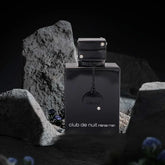 A black bottle of Luxury Men's Fragrance: Night Club Wild Black Edition 105ML by Showlu Well Nation Store sits among gray rocks, with small blue flowers visible in the dim background.