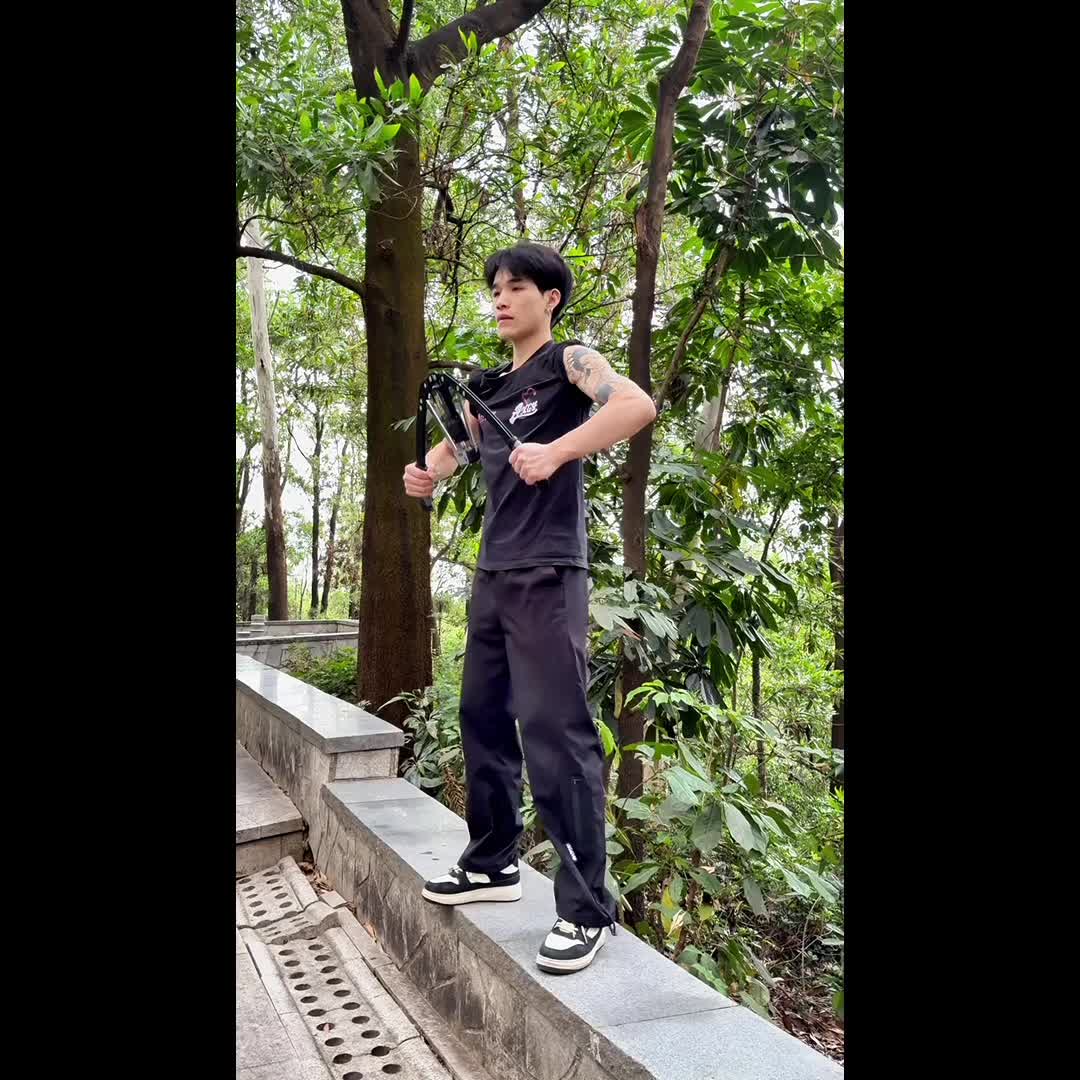 Dressed in black, a person exercises with the Versatile Hydraulic Chest Expansion Bar (22-440Lbs) on a stone forest path in Guangdong, surrounded by trees and greenery.