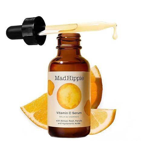 A dropper bottle of Mad Hippie Vitamin C Serum for Face with Hyaluronic Acid, Ferulic Acid, and Vitamin E sits with its dropper on top as serum drips out. Orange slices behind highlight the product’s anti-aging and glow-boosting benefits.