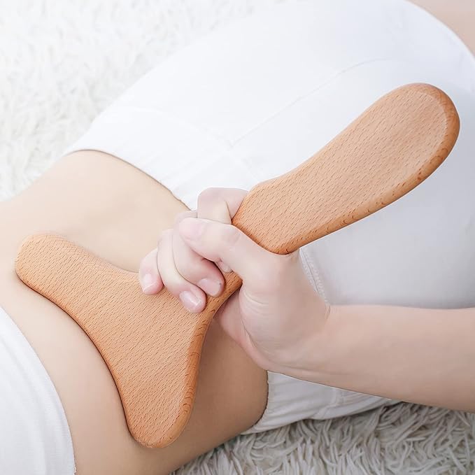 A person in white clothing uses the Wood Therapy Massage Tool Wooden Lymphatic Drainage Massager Body Sculpting Tools for Maderoterapy, Gua Sha, Muscle Release (11*6 inches) on their bare abdomen while lying on a white textured surface.