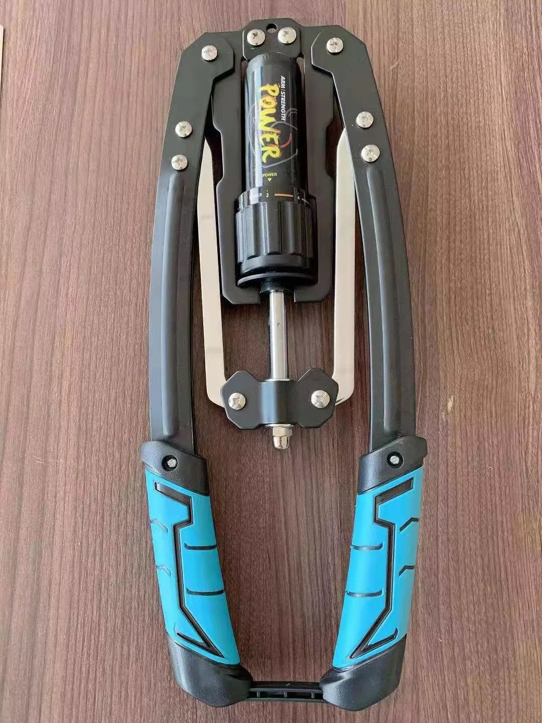 An Adjustable 22-440lbs Hydraulic Power Twister Arm Exerciser with blue and black rubber grips, metal arms, and a “Power” labeled mechanism is shown on a wooden surface, ideal for chest expander and shoulder muscle training.