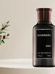 A dark bottle labeled Bharara King EDP 3.4oz, a bold woody amber men's cologne with a long-lasting luxury scent, stands on a white surface—perfect for evening wear—beside a leafy plant and neutral background; ideal holiday gift.