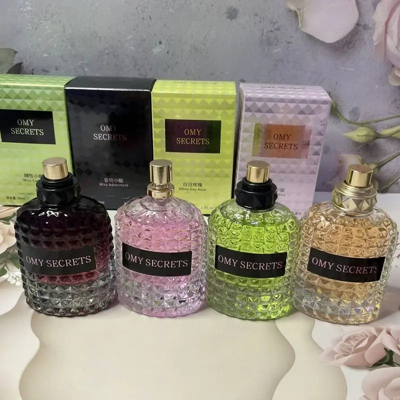 Four textured glass bottles of Miss Adventure Donna Perfume in dark purple, pink, green, and yellow stand before matching boxes on a floral-themed surface, showcasing their fresh floral-fruity scents of rose and jasmine.