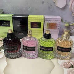 Four textured glass bottles of Miss Adventure Donna Perfume in dark purple, pink, green, and yellow stand before matching boxes on a floral-themed surface, showcasing their fresh floral-fruity scents of rose and jasmine.