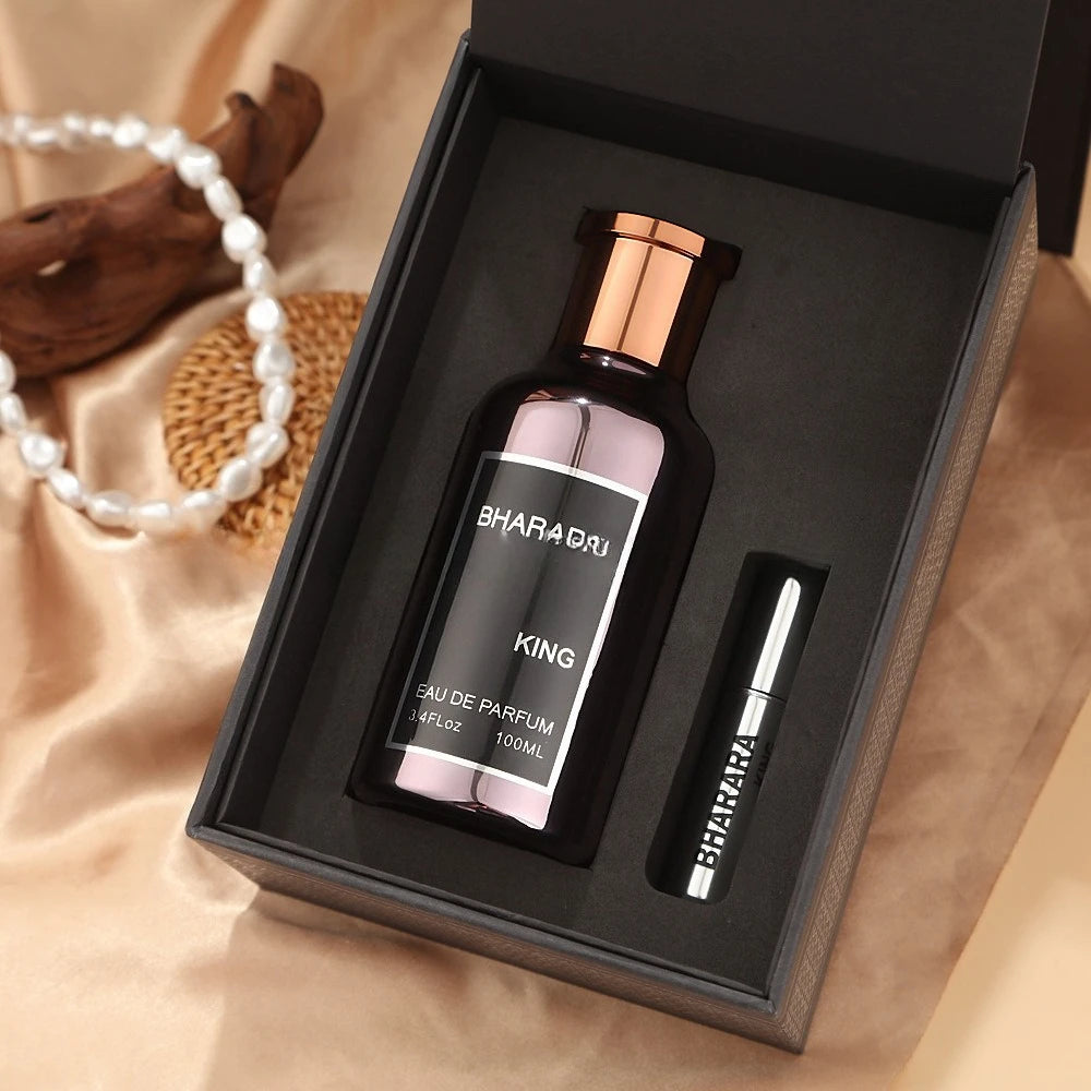 A high-end C2U Bharara King Eau De Parfum 100ML gift box for men rests on satin, featuring a large spray bottle and a travel vial. A pearl necklace and wood accent hint at its refined floral aroma.