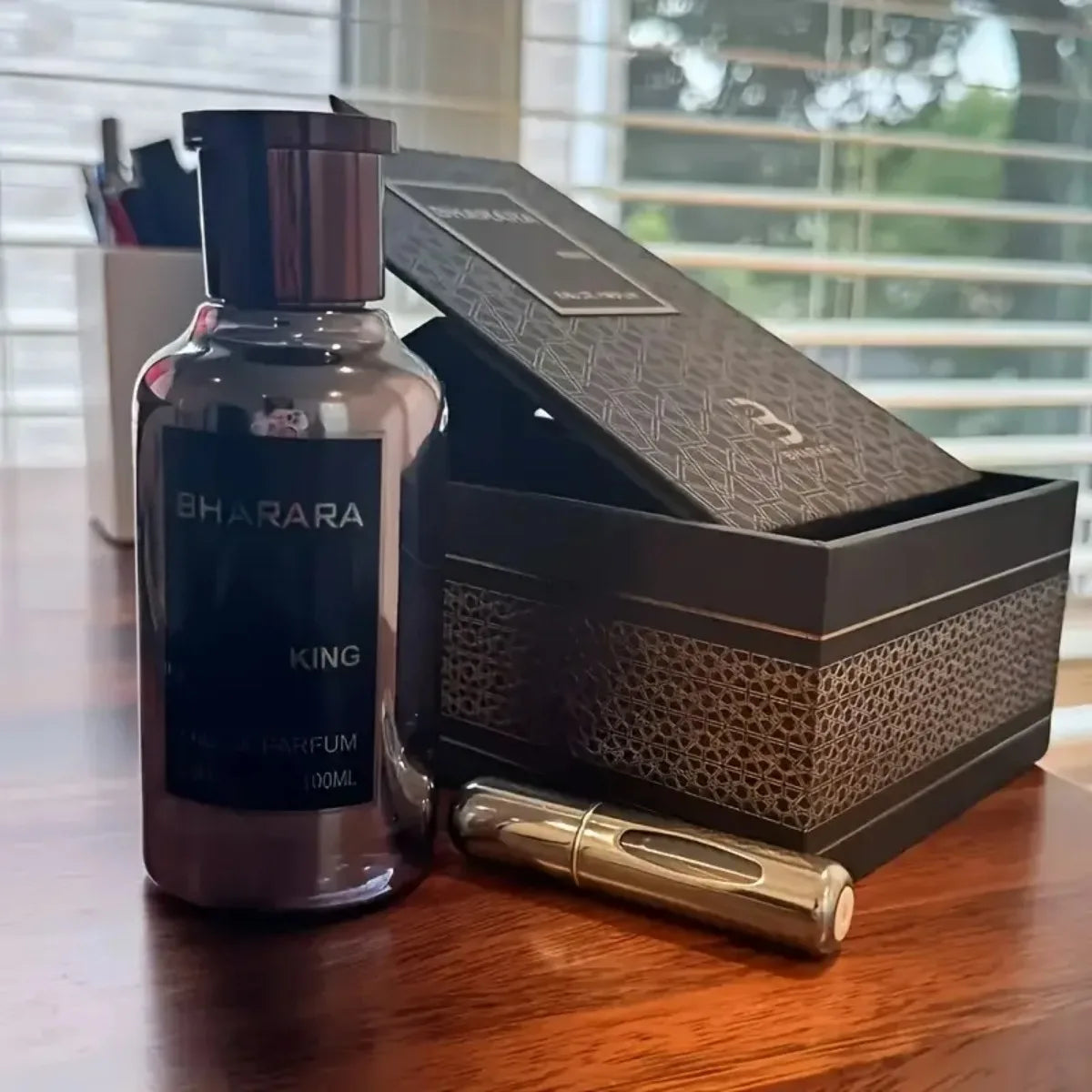 A bottle of Bharara King Eau De Parfum 100ml for Men, a citrus fruity cologne with notes of orange, amber, musk, and vanilla, sits next to a metallic spray container and an open black box on a wooden table by a window.