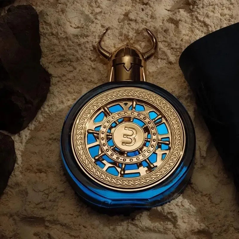 The Bharara Men's 3.4 Ounce Eau de Parfum, a round blue bottle with gold detailing and a central "3," rests on sand near dark rocks and black fabric—perfect as a holiday gift.