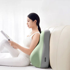A woman in white sits on a bed against a cream headboard, reading with support from the Multi-Functional Ergonomic Bed Pillow—an ergonomic memory foam wedge-shaped cushion. Natural light filters through sheer curtains at the window.