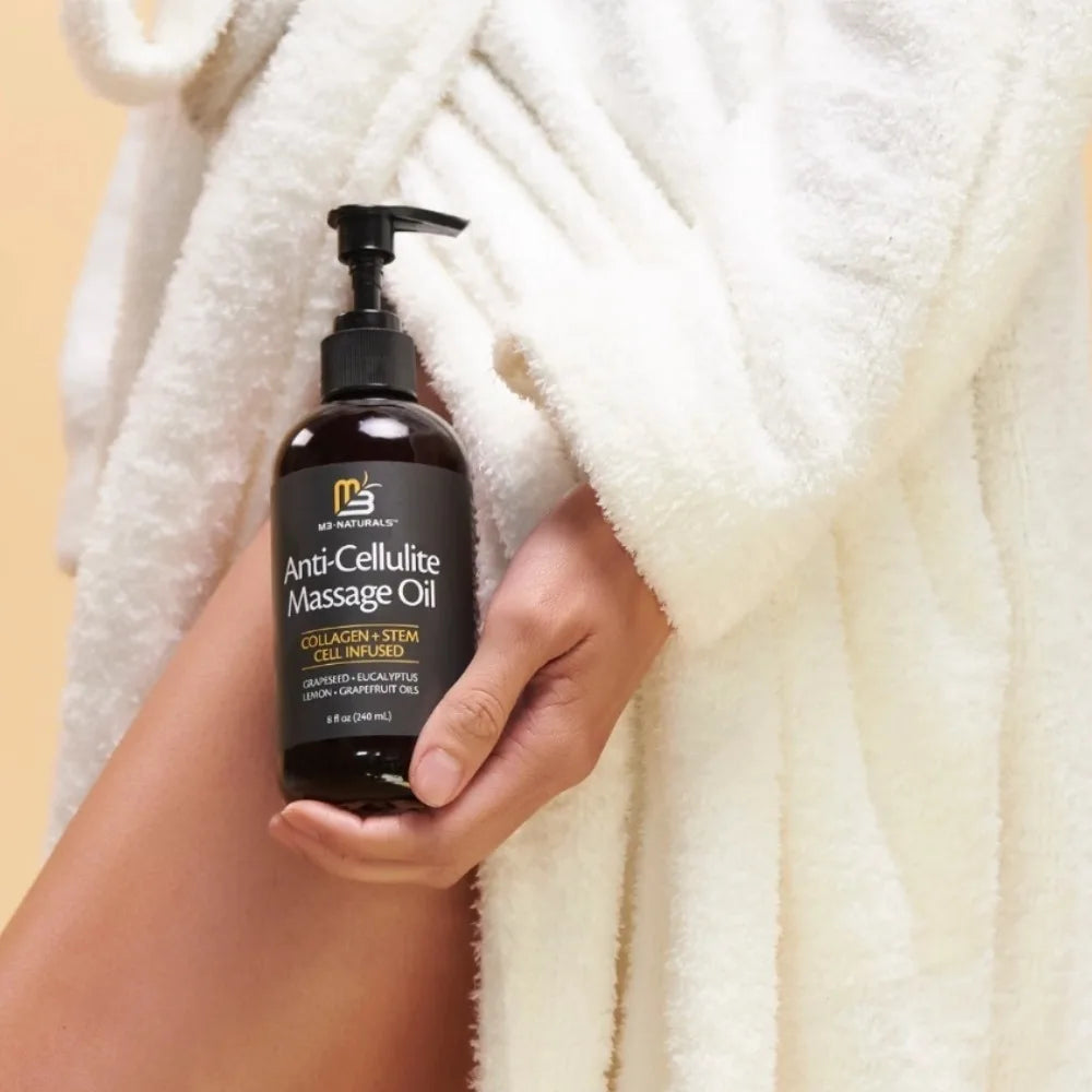A person in a white bathrobe holds a bottle of M3 Naturals Anti-Cellulite Massage Oil with grape seed and eucalyptus oils against their thigh for a moisturizing, firming treatment that improves the appearance of cellulite.