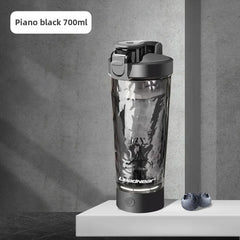 A Premium Electric Protein Shaker Bottle (700ml, piano black), USB rechargeable, stands on a gray surface beside two blueberries, set against a gray concrete background.