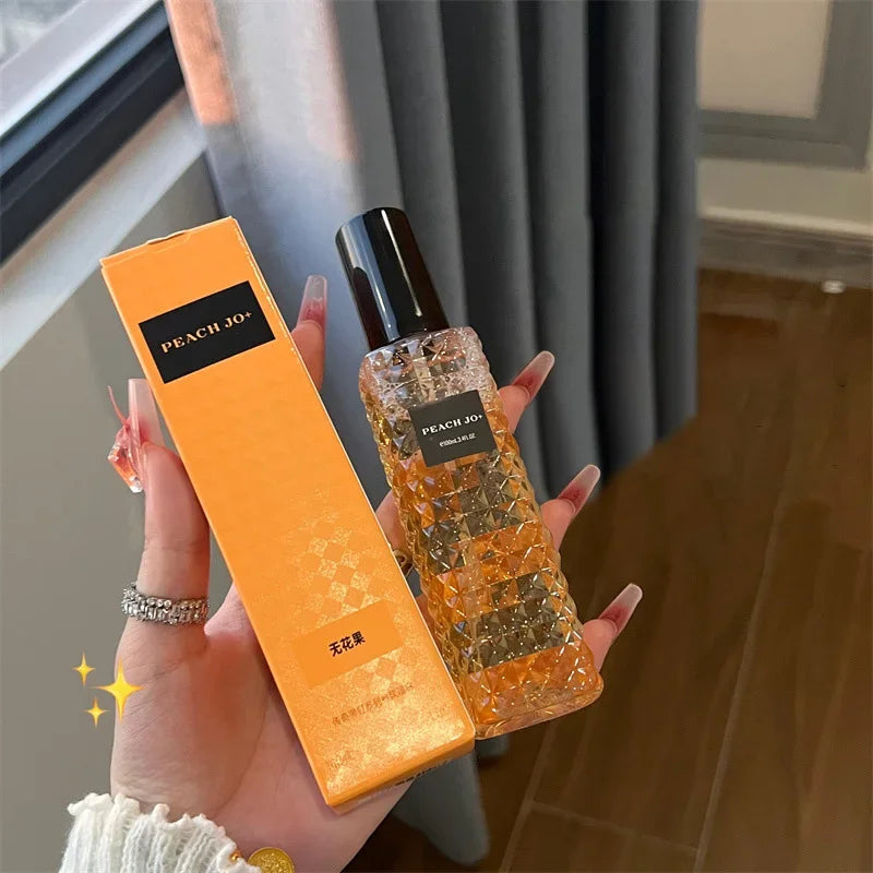 A hand holds a box and bottle labeled Rivet Series Fresh Perfume, featuring a woody, fruity, floral long-lasting light fragrance for ladies with bergamot notes. The orange set is alcohol-free. A window, curtain, and wooden floor are behind.