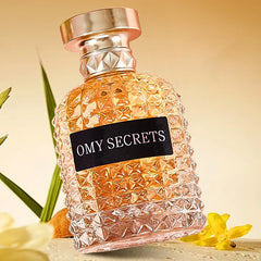 A textured glass bottle labeled Miss Adventure Donna stands upright among yellow flowers and green leaves, suggesting a fresh floral fruity scent with rose and jasmine, set against a warm, softly blurred background.