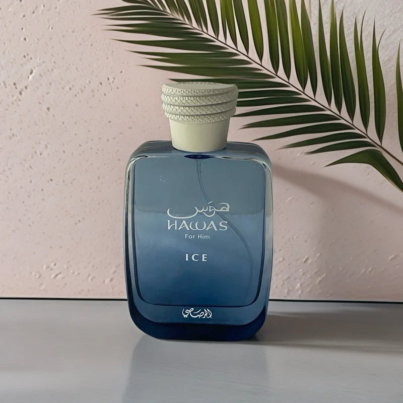 A blue bottle of RASASI Hawas Men's Ice Eau De Parfum 100ml, featuring white Arabic and English text, rests on a gray surface while a green palm leaf casts a shadow on the pale, textured wall behind it.