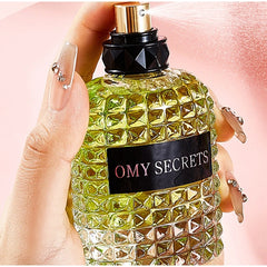 A hand with decorated nails sprays Miss Adventure Donna Perfume, a luxurious floral fruity scent with rose and jasmine, from a textured greenish-yellow bottle against a soft pink background.