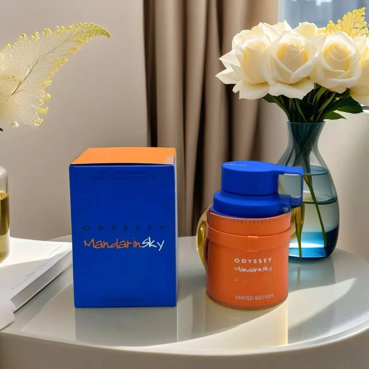 A blue and orange box labeled "Armaf Odyssey Unisex Perfume Spray, Mandarin Sky Elixir, Limited Edition, 3.4 oz" sits by its matching bottle on a round table, with white roses in a vase and beige curtains in the background.
