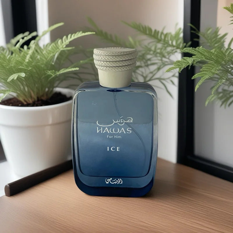 A blue glass bottle of RASASI Hawas Men's Ice Eau De Parfum 100ml stands on a wooden surface beside a white potted plant with green leaves, with a window in the background. The label highlights this vibrant men's fragrance.