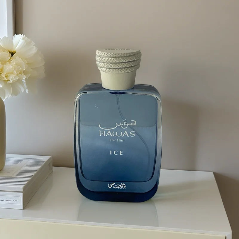 A blue glass bottle of RASASI Hawas Men's Ice Eau De Parfum 100ml, featuring white Arabic and English text, rests on a white surface near a white flower and stacked books—an elegant fragrance choice for men against a beige backdrop.