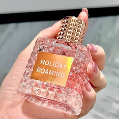 A hand holds a pink, textured Rivet Series Fresh Perfume bottle with a gold-studded cap. This long-lasting eau de toilette offers a woody, fruity, and floral fragrance with bergamot notes. Background is blurred in neutral tones.