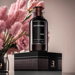 A dark bottle of C2U Bharara King Eau De Parfum 100ML for Men rests on its patterned black gift box, with soft pink flowers in the background, highlighting this high-end 3.4oz spray perfume’s elegant presentation.