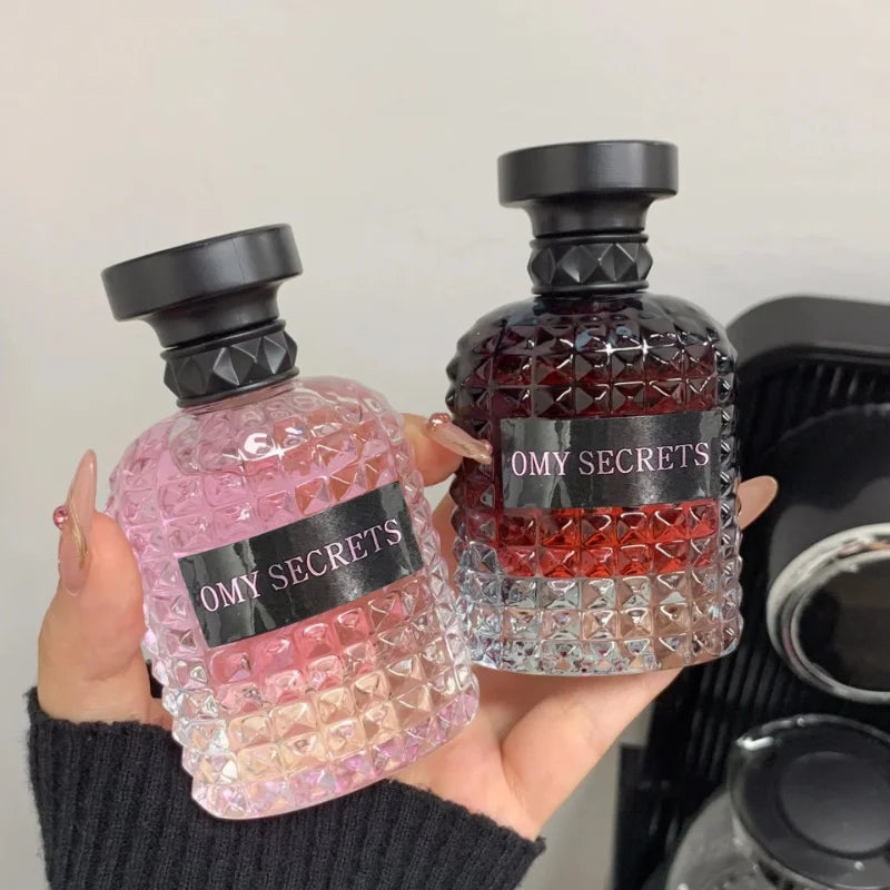 A hand holds two textured glass bottles labeled Rivet Series Fresh Perfume, one pink, one fading from dark red to clear. Both feature black caps and offer a long-lasting woody, fruity, floral scent with bergamot in an alcohol-free formula.