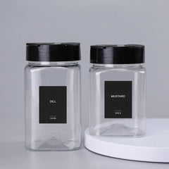 Two clear square spice jars with black lids are labeled "Dill (herb)" and "Mustard (spice)" using 32Pcs Waterproof Rectangle Spice Bottle Labels Self Adhesive Stickers, sitting side by side on a gray surface.