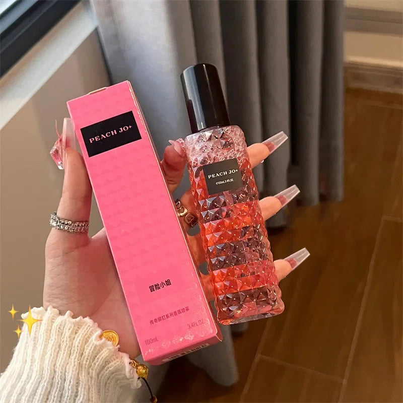 A hand with long, decorated nails holds a textured pink bottle labeled "Rivet Series Fresh Perfume"—a woody, fruity, floral ladies’ fragrance—and its matching pink box. The background features a wooden floor and gray curtain.