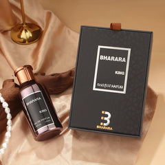 A bottle of C2U Bharara King Eau De Parfum 100ML for Men, a high-end spray perfume, rests beside its sealed black and gold gift box on beige fabric, accompanied by a pearl necklace, gold stand, and wooden accent.