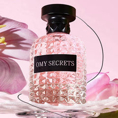 A textured glass bottle labeled Miss Adventure Donna sits on a reflective surface amid pink flowers and petals, featuring a lasting fresh rose and jasmine floral fruity scent, against a soft pink backdrop.