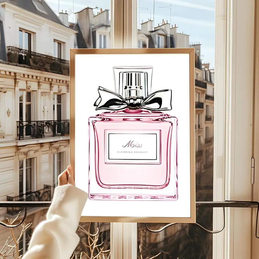 A person holds up a framed "Perfume Bottle Prints Nordic Poster Wall Art" by a window with Parisian buildings visible in the background.