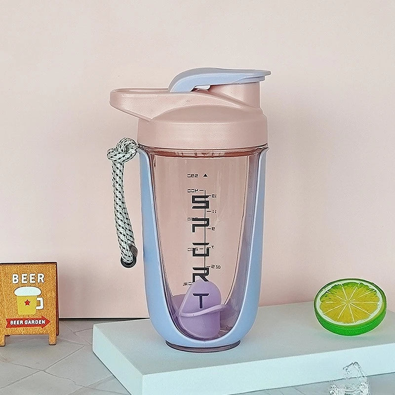 A pink and purple Premium Protein Powder Shaker (590ml/20oz), BPA-free Tritan with flip-top lid, rope handle, and plastic ball, stands on a white platform beside a fake lemon slice, "BEER" sign, and ice cube on marble. Made in China.