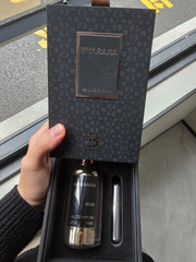 A hand holds an open black box with a geometric pattern, containing a 100ML Brand Man Musk Perfume—Long Lasting Citrus Floral Light Fragrance—and a small silver spray vial, by a window overlooking the street.