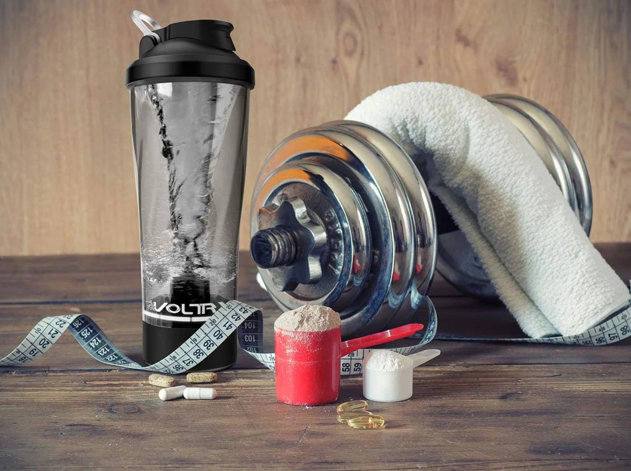 The Premium Electric Protein Shaker Bottle USB Rechargeable Portable Mixer Cup for Fitness Protein Shakes is arranged on a wooden surface alongside a metal dumbbell, white towel, measuring tape, supplements, scoop, and capsules.