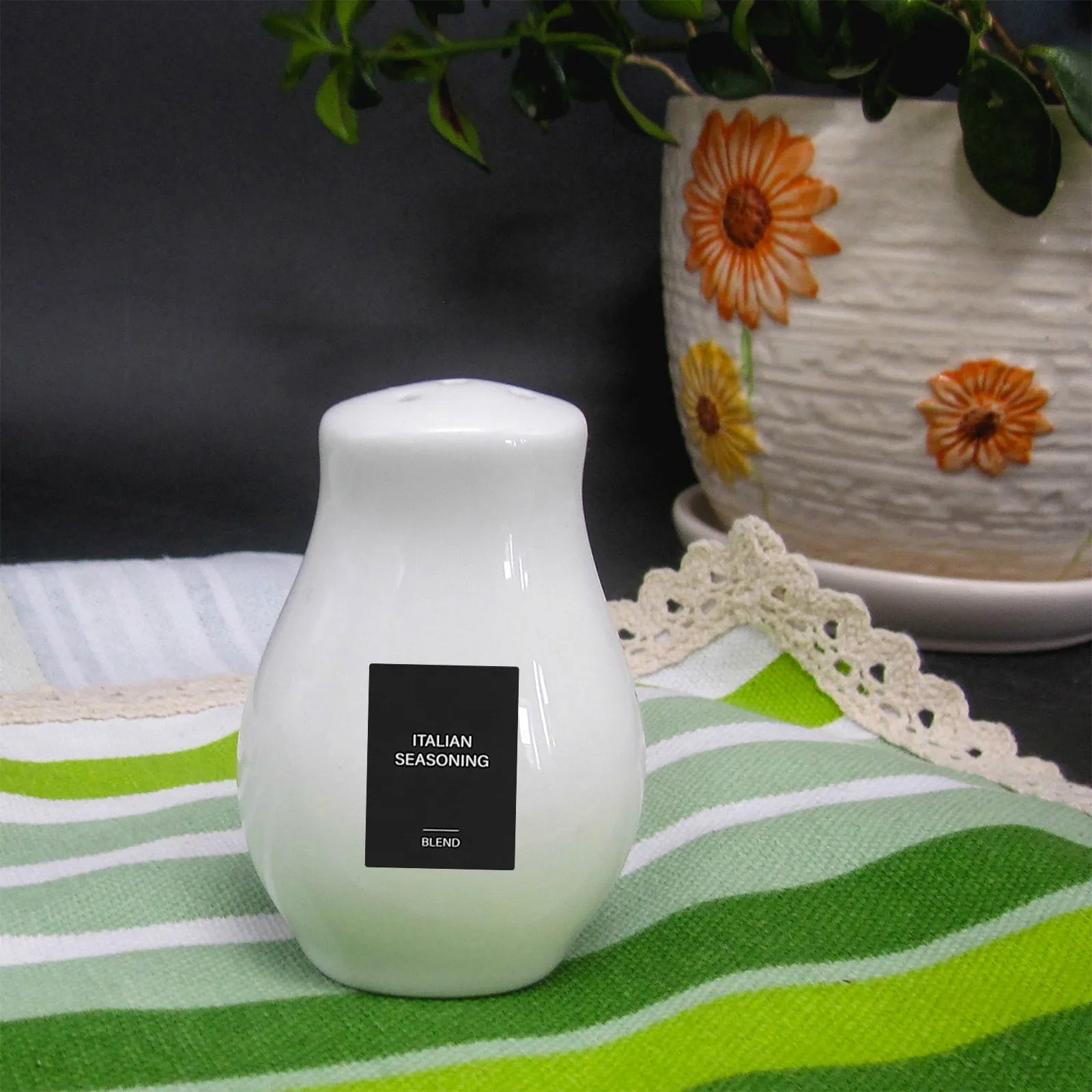 A white ceramic shaker with a 32Pcs Waterproof Rectangle Spice Bottle Label for Italian Seasoning Blend sits on a green striped cloth, next to a flower-patterned plant pot and green leaves in the background.