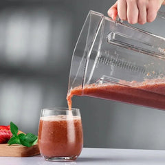A hand pours a reddish-brown smoothie from the For SILVER CREST Blender Spare Jug (2L, transparent) into a glass. Strawberries, mint leaves, and the blender jug are on a cutting board in the background on a gray surface.
