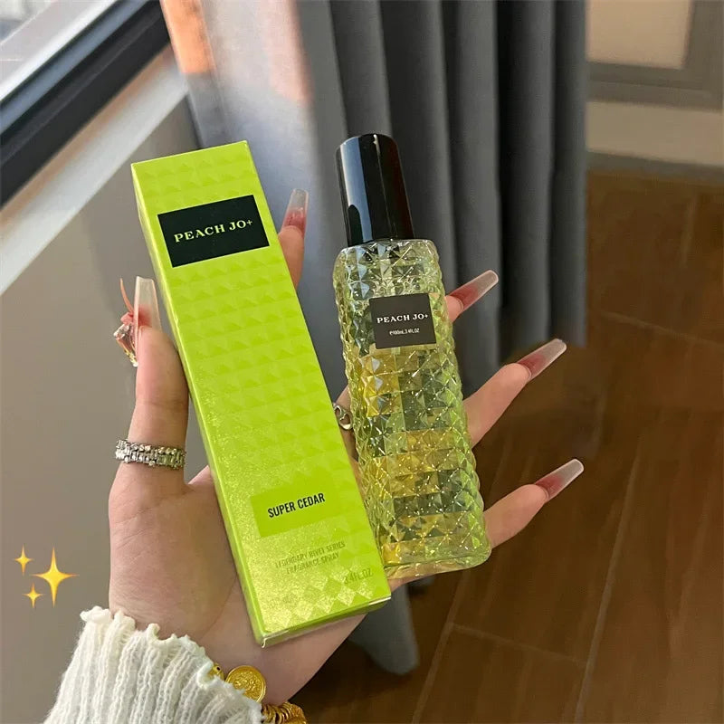 A manicured hand holds a textured glass bottle labeled Rivet Series Fresh Perfume, a woody fruity floral, alcohol-free fragrance. Beside it is a matching green box. A curtain and window are in the background.