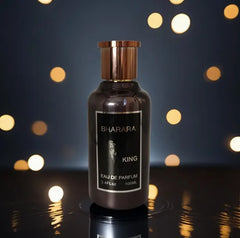 A 100ml bottle of Bharara King Eau De Parfum for men, with a gold cap and label, stands upright on a reflective surface against warm bokeh lights, highlighting its sleek, luxurious design popular in Mainland China.