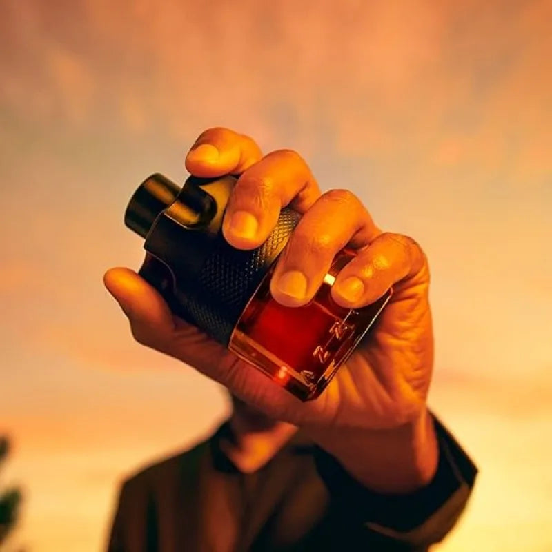 A hand holds an Azzaro The Most Wanted Parfum Intense Mens Cologne 100ML bottle toward the camera at sunset with a warm orange sky, focusing on the bottle and hand while the face is blurred in the background.