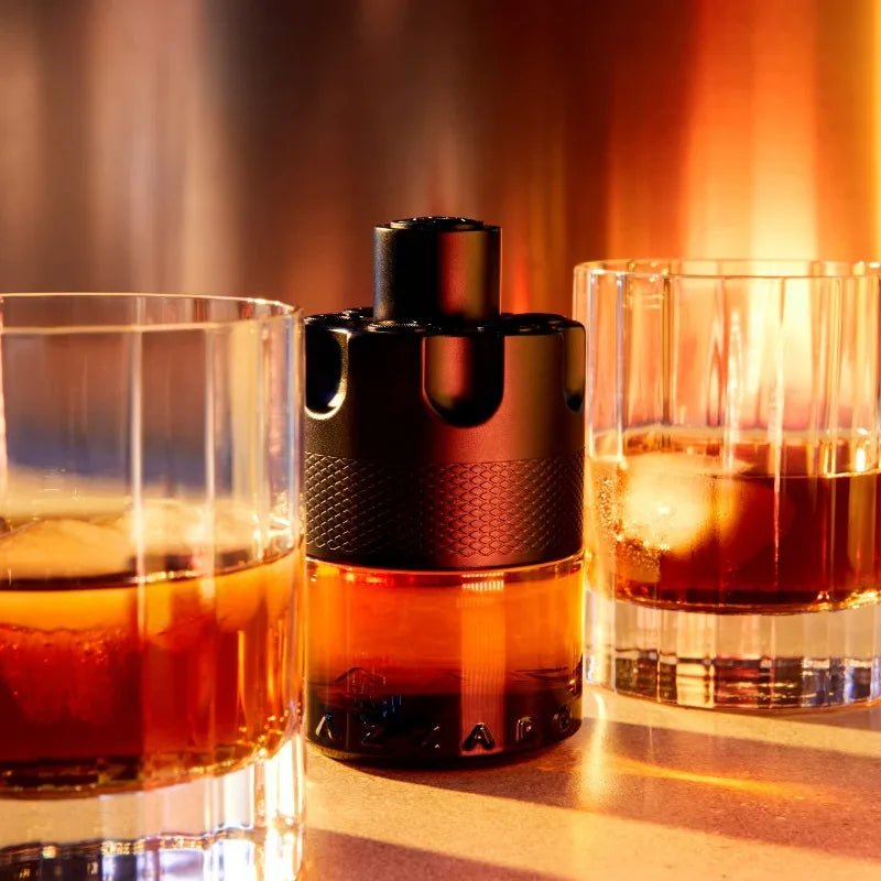 A close-up of two whiskey glasses with amber liquid and ice, with a 100ml Azzaro Wanted Series Eau De Parfum Spray bottle between them, set against a warm, glowing background.