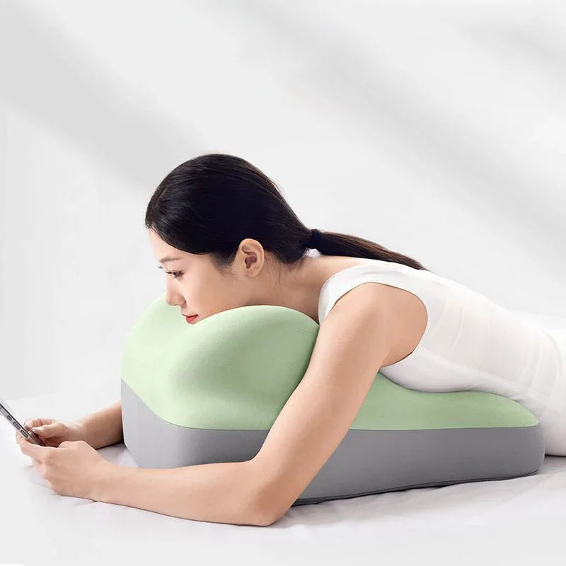 A woman in a white outfit lies on her stomach, using the Multi-Functional Ergonomic Bed Pillow—a memory foam wedge-shaped cushion with a 100% cotton cover—to support her chest and arms while looking at her phone.