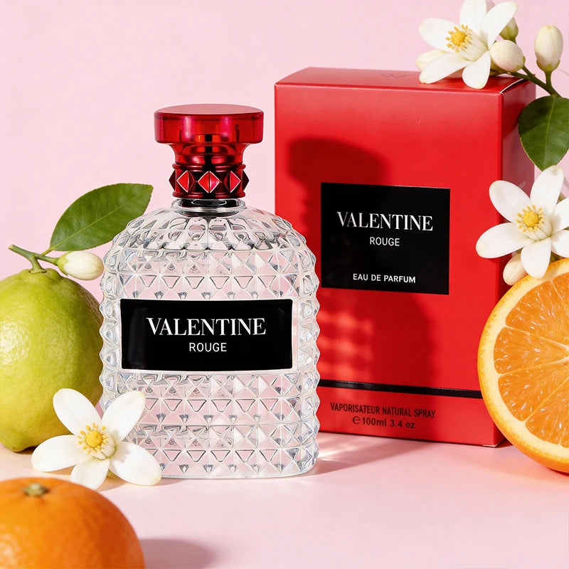 A clear, textured perfume bottle labeled 3.4 Fl Oz Men's and Women's Eau De Parfum stands by a matching red box, surrounded by mandarin oranges, orange blossoms, and bergamot—evoking a fresh holiday scent against a light pink background.