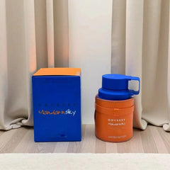 An Armaf Odyssey Unisex Perfume Spray, Mandarin Sky Elixir Limited Edition (3.4 oz) in an orange container with blue accents sits beside its blue and orange box on a wooden floor in front of beige curtains.